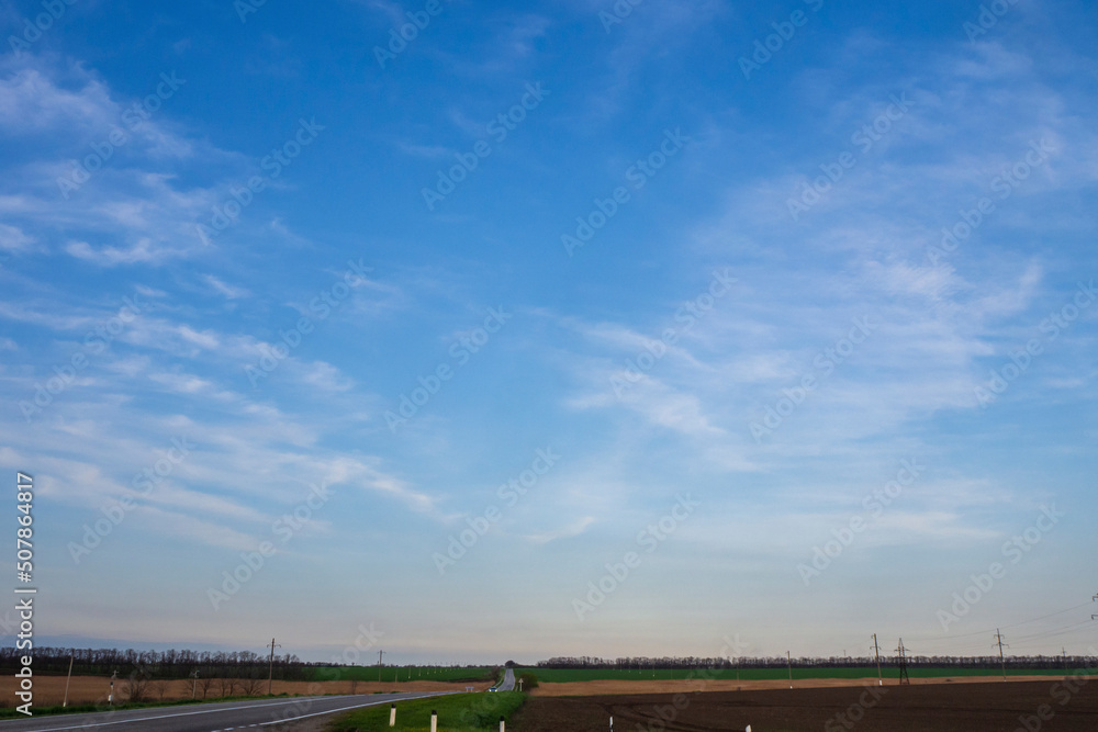 Blue sky with soft clouds over an asphalt road stretching into the distance between agricultural fields. Car travel Blue sky with soft clouds over an asphalt road stretching into the distance between agricultural fields. Car travel