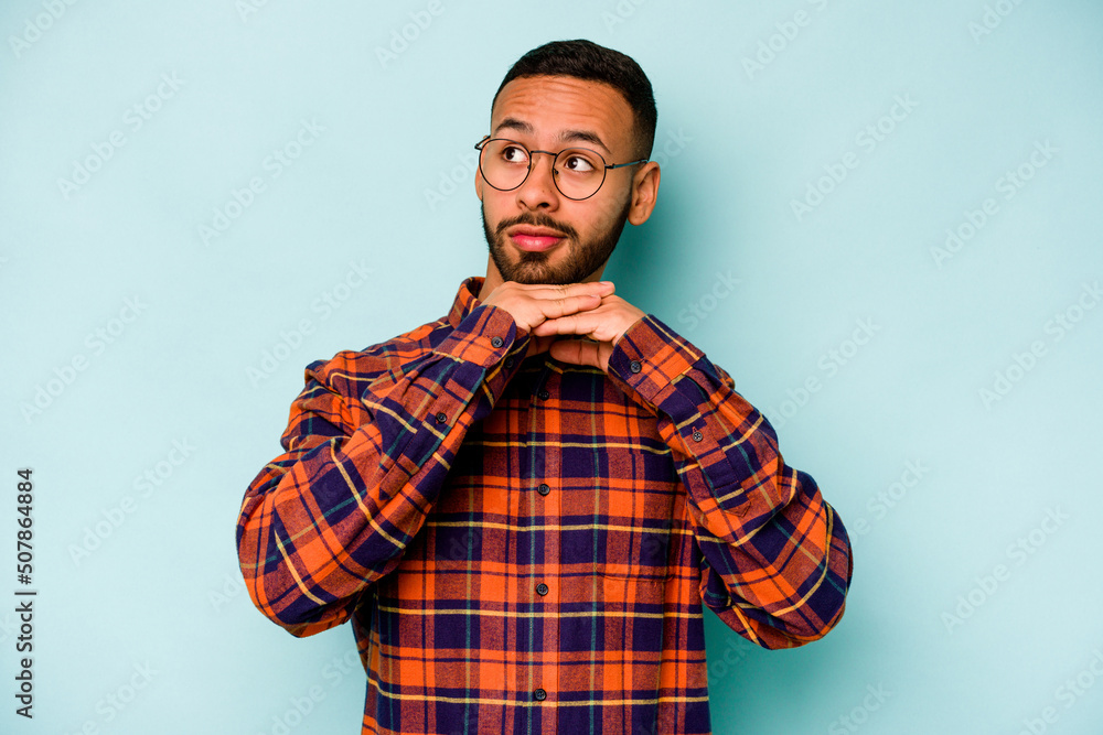 Young hispanic man isolated on blue background keeps hands under chin, is looking happily aside. Young hispanic man isolated on blue background keeps hands under chin, is looking happily aside.