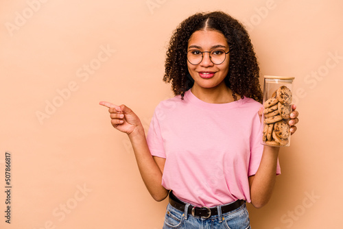 Canvastavla Young African american woman holding a cookies jar isolated on beige background smiling and pointing aside, showing something at blank space
