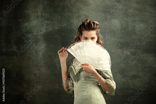 Vintage portrait of young beautiful girl in gray dress of medieval style with fan isolated on dark background. Comparison of eras concept, flemish style. Art, beauty