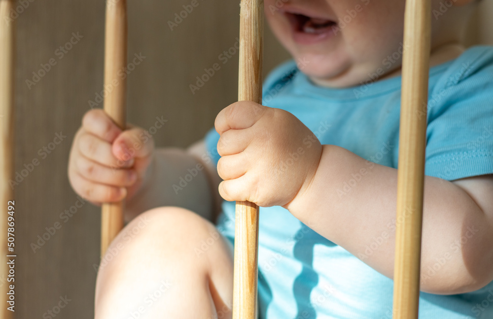 cute little baby boy is crying in the crib, holding the wooden bars