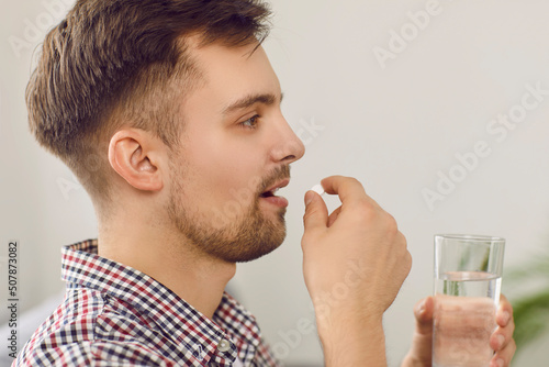 Young man takes medication prescribed by his physician. Side profile closeup view of a happy handsome Caucasian man taking a pill and drinking a glass of water. Health, medicine, treatment concept