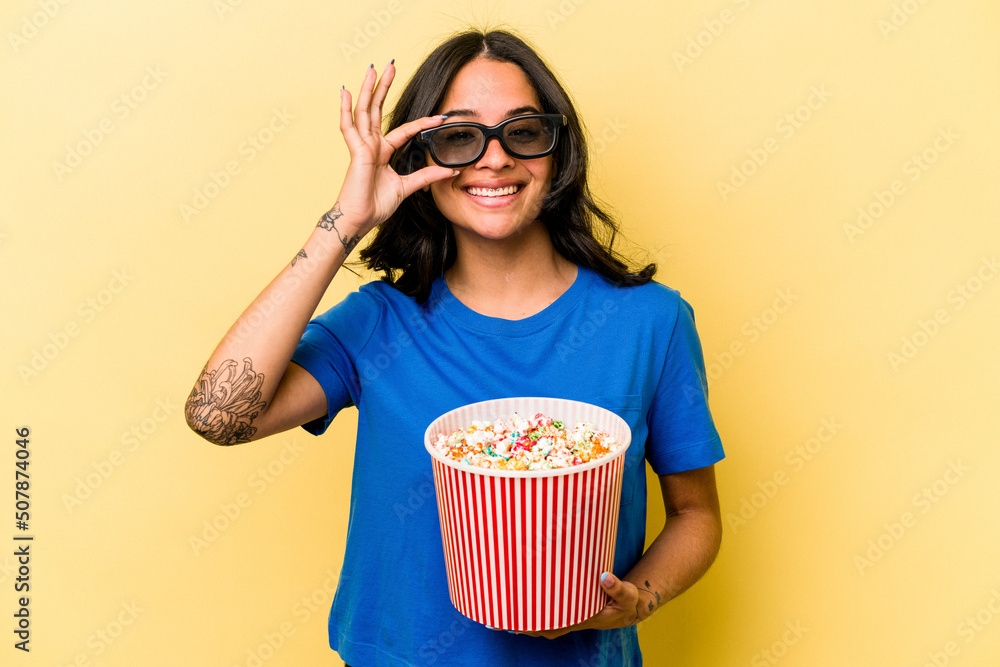 Young hispanic woman holding popcorn isolated on yellow background excited keeping ok gesture on eye.