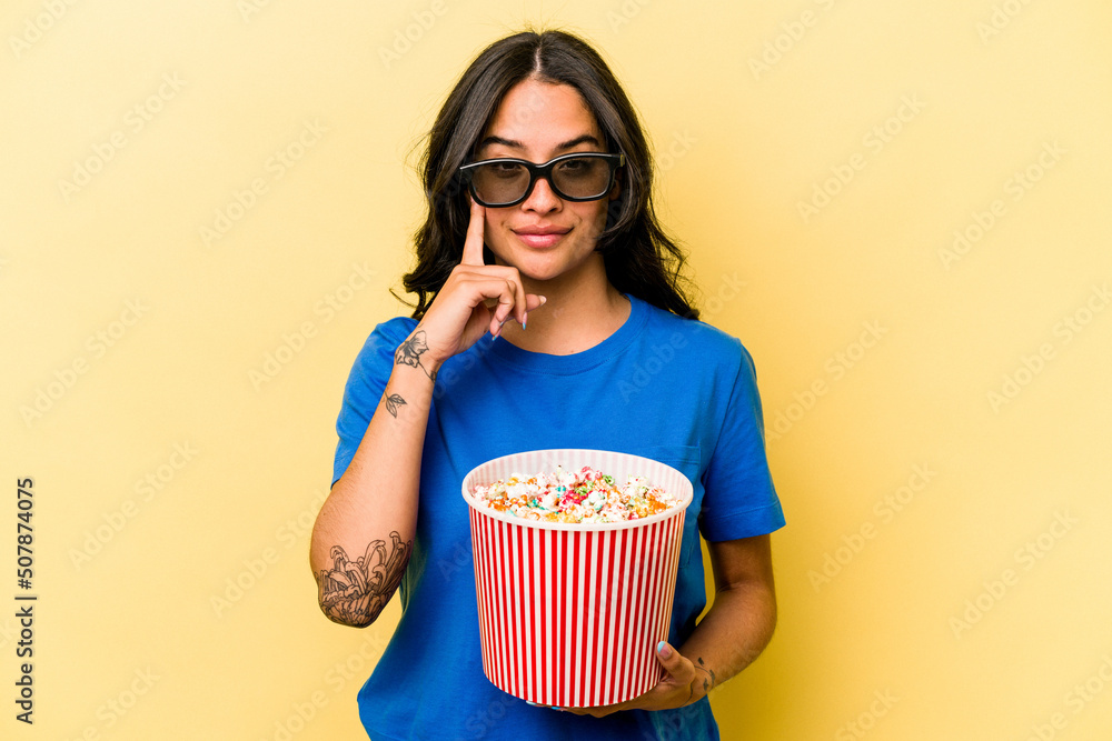 Young hispanic woman holding popcorn isolated on yellow background pointing temple with finger, thinking, focused on a task.