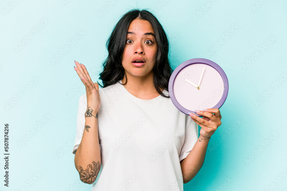 Young hispanic woman holding a clock isolated on blue background surprised and shocked. Young hispanic woman holding a clock isolated on blue background surprised and shocked.