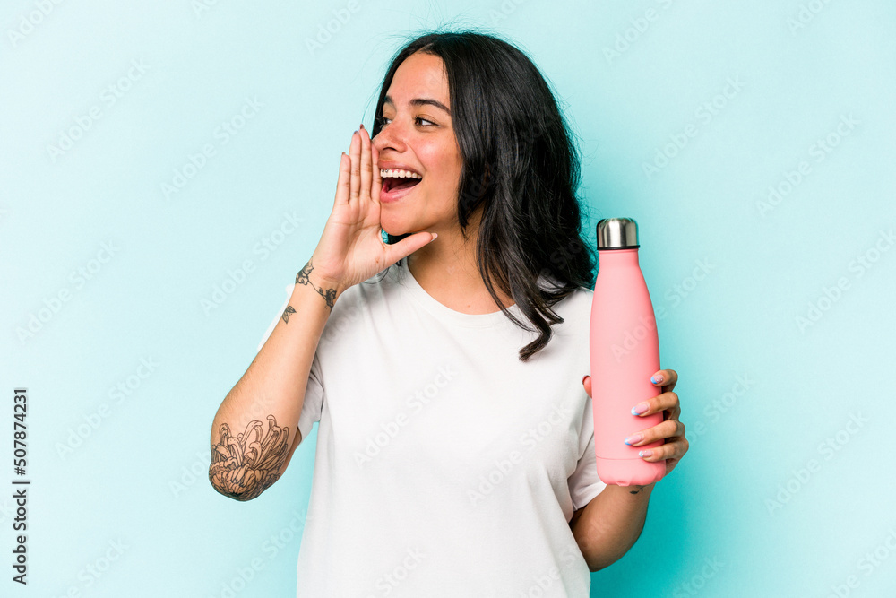 Young hispanic woman holding thermo isolated on blue background shouting and holding palm near opened mouth. Young hispanic woman holding thermo isolated on blue background shouting and holding palm near opened mouth.