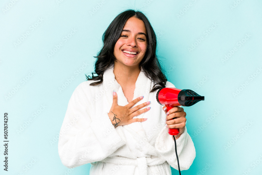Young hispanic woman holding dryer isolated on blue background laughs out loudly keeping hand on chest. Young hispanic woman holding dryer isolated on blue background laughs out loudly keeping hand on chest.