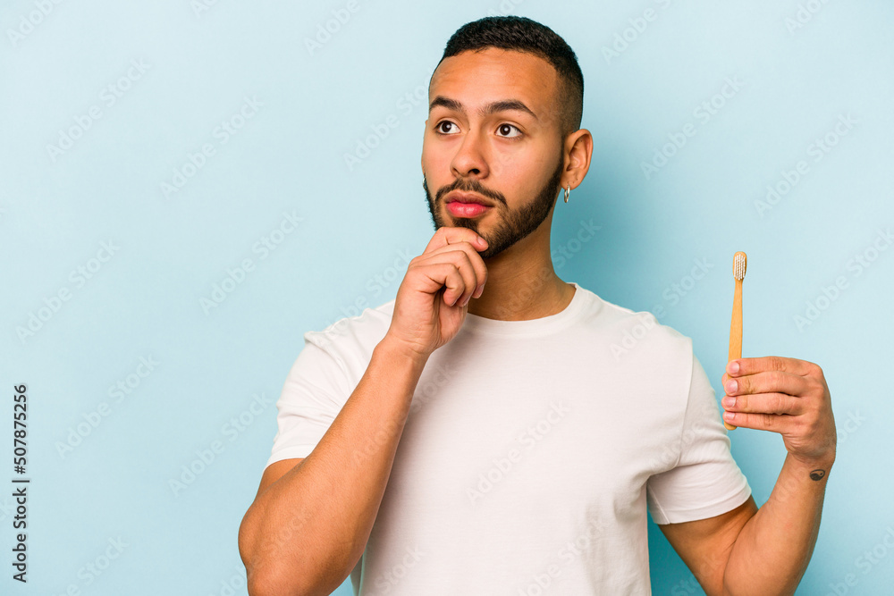 Young hispanic man brushing teeth isolated on blue background looking sideways with doubtful and skeptical expression.