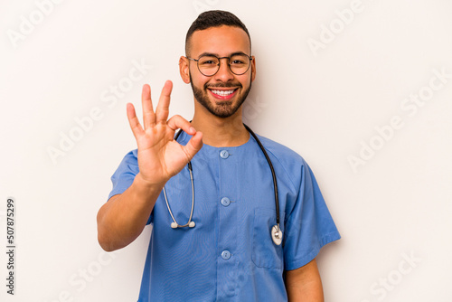 Young hispanic nurse man isolated on white background cheerful and confident showing ok gesture. © Asier