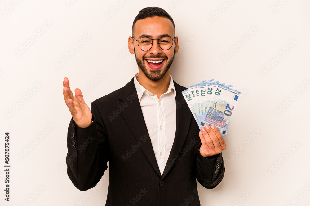 Young business hispanic man holding banknotes isolated on white background receiving a pleasant surprise, excited and raising hands. Young business hispanic man holding banknotes isolated on white background receiving a pleasant surprise, excited and raising hands.