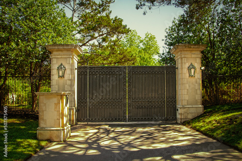 Closed impressive wrought iron luxury  security gate with key pad for private residential estate in leafy neighborhood with green trees and shadows on drive