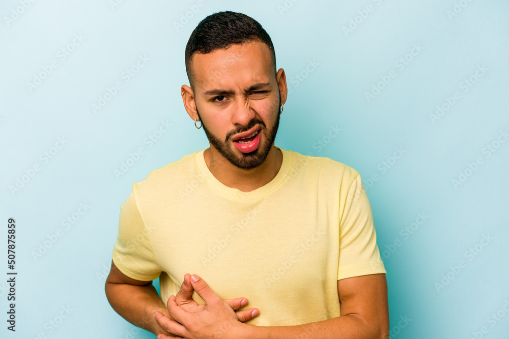 Young hispanic man isolated on blue background having a liver pain, stomach ache. Young hispanic man isolated on blue background having a liver pain, stomach ache.