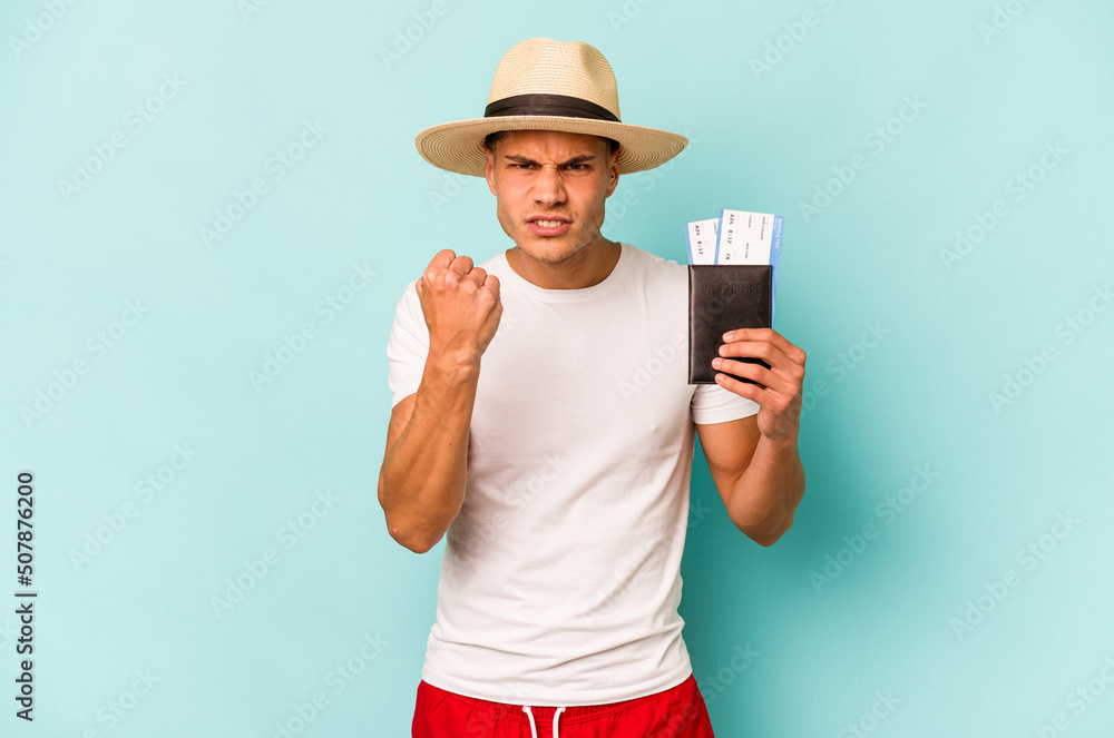 Young caucasian traveler man holding passport isolated on blue background showing fist to camera, aggressive facial expression. Young caucasian traveler man holding passport isolated on blue background showing fist to camera, aggressive facial expression.