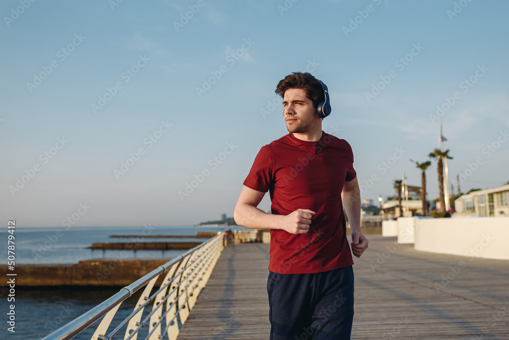 Young strong sporty athletic fit sportsman man in sports clothes heapdhones listen music look aside warm up training at sunrise sun dawn over sea beach outdoor on pier seaside in summer day morning Young strong sporty athletic fit sportsman man in sports clothes heapdhones listen music look aside warm up training at sunrise sun dawn over sea beach outdoor on pier seaside in summer day morning
