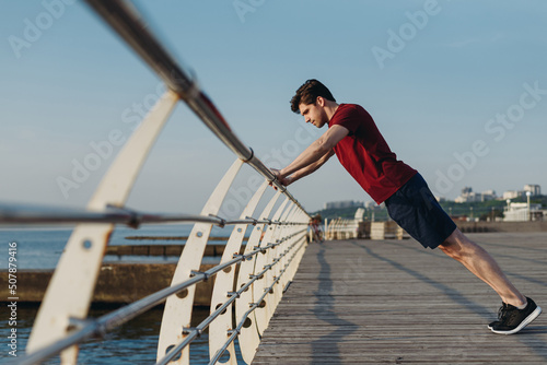 Full body side view young strong sporty toned fit sportsman man in sports clothes do push-ups exercises on railing warm up train at sunrise sun dawn over sea beach outdoor on pier seaside in morning.
