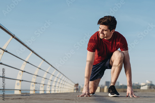 Full size young strong sporty toned fit sportsman man 20s wear sports clothes stand at low start going to run warm up training at sunrise sun dawn over sea beach outdoor on pier seaside in morning