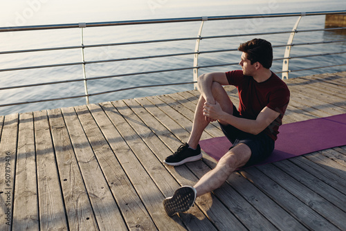 Full length young strong sporty athletic toned fit sportsman man in sports clothes sitting on yoga mat warm up train at sunrise sun dawn over sea beach outdoor on pier seaside in summer day morning