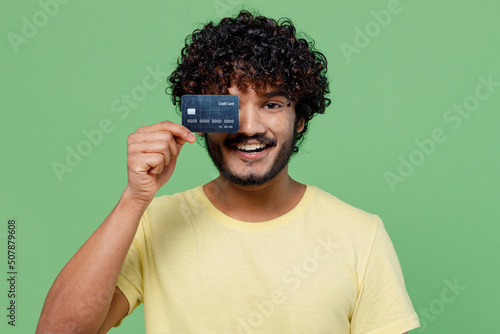 Young smiling happy satisfied cheerful Indian man 20s in basic yellow t-shirt hold in hand cover eye credit bank card isolated on plain pastel light green background studio. People lifestyle concept.
