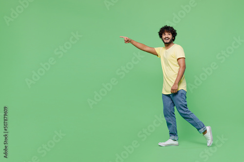 Full body young smiling happy Indian man 20s wearing basic yellow t-shirt walk going strolling point index finger aside isolated on plain pastel light green background studio People lifestyle concept.