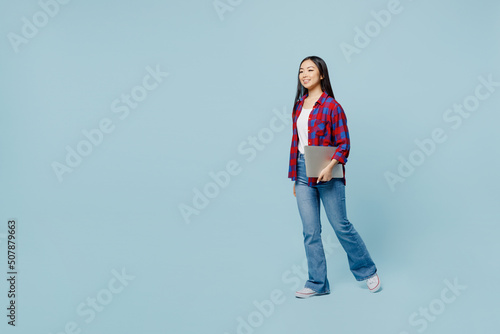 Full body young smiling fun woman of Asian ethnicity wear checkered shirt hold closed laptop pc computer walking going isolated on plain pastel light blue color background. People lifestyle concept.