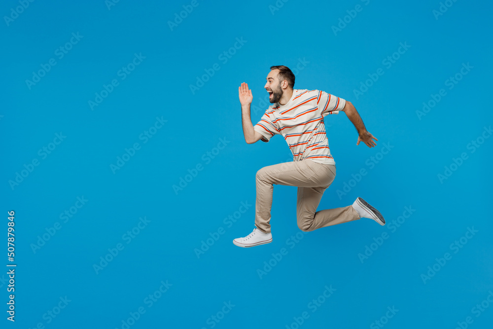 Full body side profile view young excited happy fun caucasian man 20s wearing orange striped t-shirt run fast hurry upisolated on plain blue color background studio portrait. People lifestyle concept.