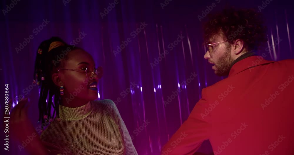 Young man in red jacket and silver chain dances face to face with young woman in fishnet top and sunglasses. Girl and guy spend evening together in colorful flashes of night club. Young man in red jacket and silver chain dances face to face with young woman in fishnet top and sunglasses. Girl and guy spend evening together in colorful flashes of night club.