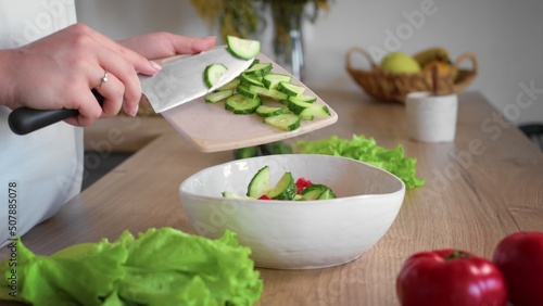 Close-up of an unrecognizable woman's hand cutting a cucumber with a kitchen knife. A woman transfers a sliced cucumber to a bowl. Cooking in the kitchen.