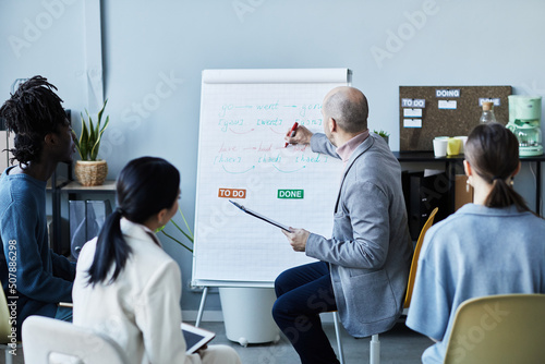 Portrait of male teacher writing on whiteboard while giving English lesson to group of people sitting in circle during seminar in office