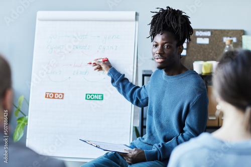 Portrait of young black man teaching English class in office to group of people and pointing at whiteboard with grammar rules