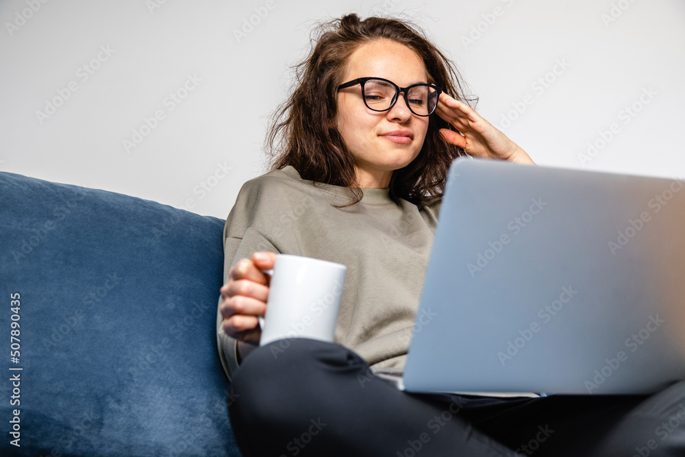 Young brunette woman using laptop and drinking coffee while sitting on sofa at home. Young brunette woman using laptop and drinking coffee while sitting on sofa at home.