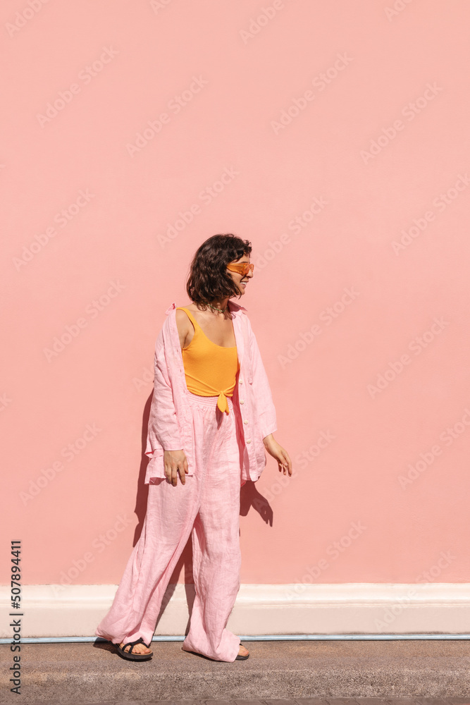Full-length, cheerful young caucasian woman stands near pink-colored street wall. Brunette is wearing casual summer clothes in fine weather. Concept of enjoying weekend, vacation Full-length, cheerful young caucasian woman stands near pink-colored street wall. Brunette is wearing casual summer clothes in fine weather. Concept of enjoying weekend, vacation