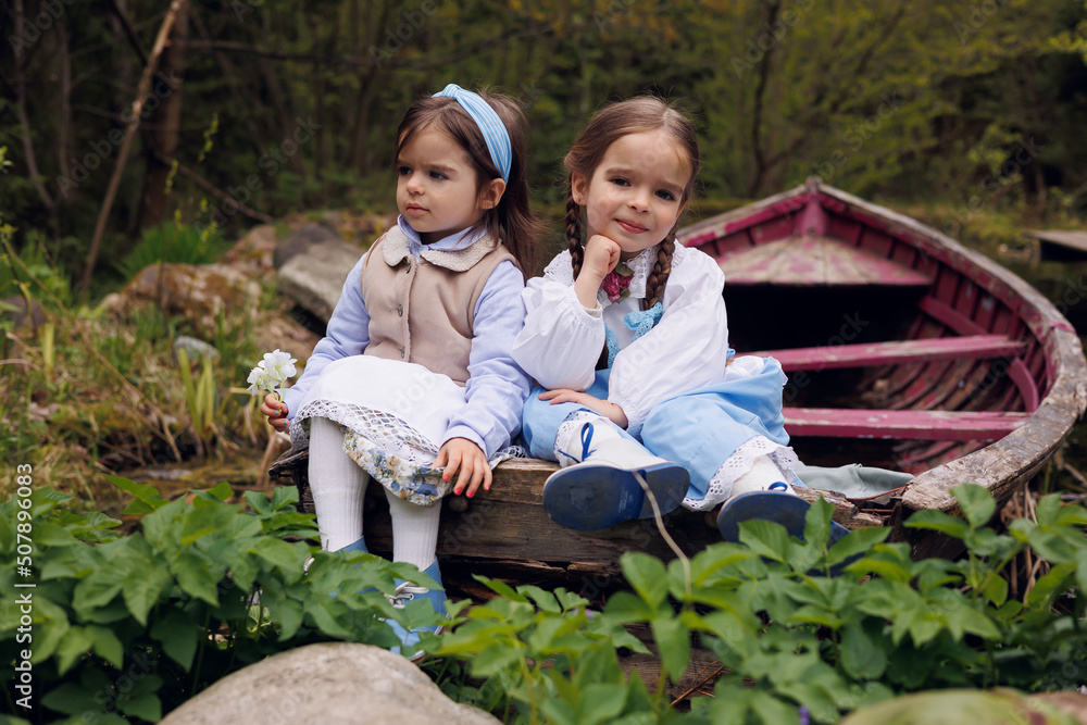 Two little girls of 3 and 5 sitting in old wooden pink boat in village. Little baby sisters in retro vintage dress smiling and thoughtful