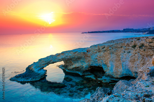 Beautiful Arch in the rocks by the sea in Ayia Napa. The bridge of lovers on the background of a bright sunny pink sunset. Travel to Cyprus tourism sightseeing islands. photo