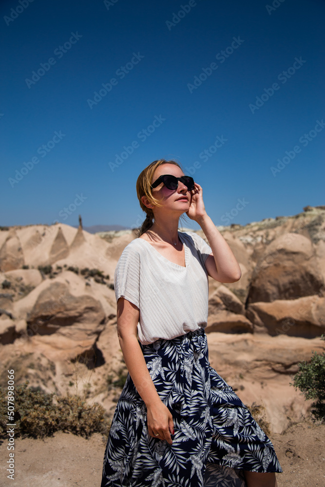 Fototapeta premium Young happy woman traveler enjoying a vacation in desert Cappadocia Turkey. Tourist girl in sunglasses and dress outdoor portrait. Sunny female portrait in the desert.