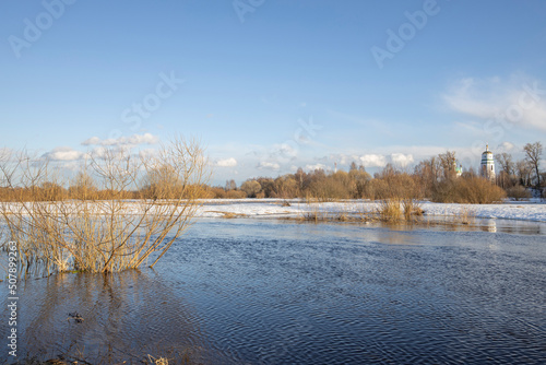March sunny day by the river. A picturesque landscape, early spring, a river with snow-covered banks, dry grass and bushes. Church in the background. The first thaws, the snow is melting © Sergei