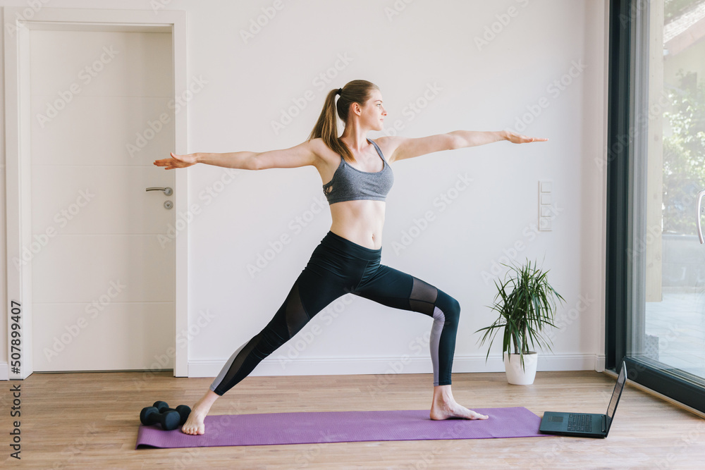 Young woman practices yoga class at home. The girl athlete stands in the pose of a warrior. Education and training online. Poses and concept of yoga. Young woman practices yoga class at home. The girl athlete stands in the pose of a warrior. Education and training online. Poses and concept of yoga.