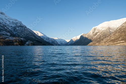 Dark blue water with small waves in a Norwegian fjord where the gray stone mountains have white snow on which the sun shines