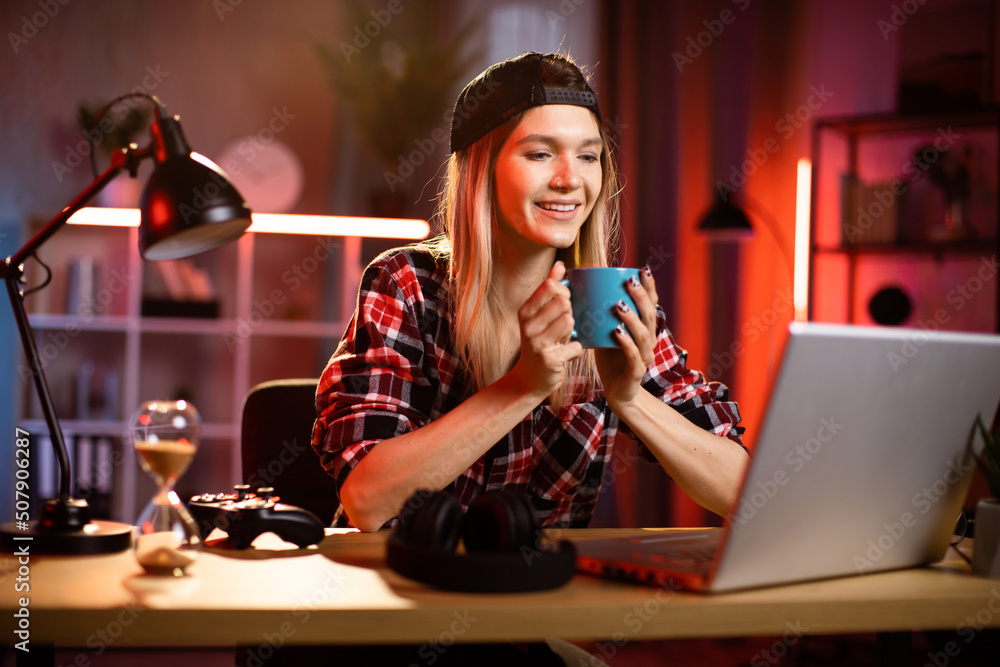 Likable young blond Caucasian woman sitting at desk with laptop and hot drink in her hands. Pleasant female in checkered shir using portable computer for work at dark room at home drinking coffee. Likable young blond Caucasian woman sitting at desk with laptop and hot drink in her hands. Pleasant female in checkered shir using portable computer for work at dark room at home drinking coffee.