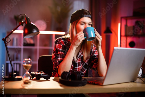 Likable young blond Caucasian woman sitting at desk with laptop and hot drink in her hands. Pleasant female in checkered shir using portable computer for work at dark room at home drinking coffee. photo