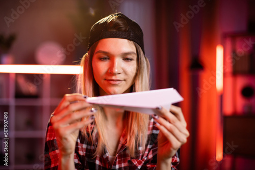Attractive caucasian woman in checkered shirt playing with paper plane during evening time. Young female freelancer sitting at desk and distracted from work at home.