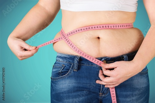 Unrecognizable fat plump plus-size overweight woman standing, showing excess naked belly, measuring waist with tape. Dieting, unhealthy food, obesity.