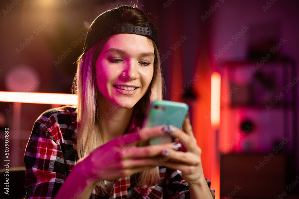 Pretty young woman in checkered shirt sitting at desk with modern smartphone in hands. Beautiful happy lady surfing internet on cell phone during evening time at home.