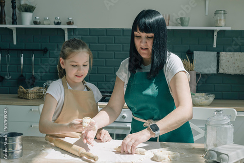 Mom teaching daughter to preparing dough on kitchen table. Young girl helping mother on kitchen.