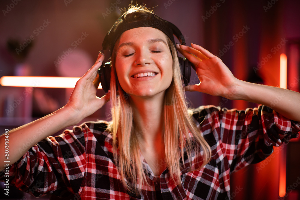 Naklejka premium Pretty woman and listening music in holding headphones in hands. Young lady in checkered shirt sitting at desk with wireless laptop. Relaxation during evening work at home.