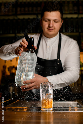Obraz na plátně male bartender holds a siphon and splashes on glass with a cold cocktail