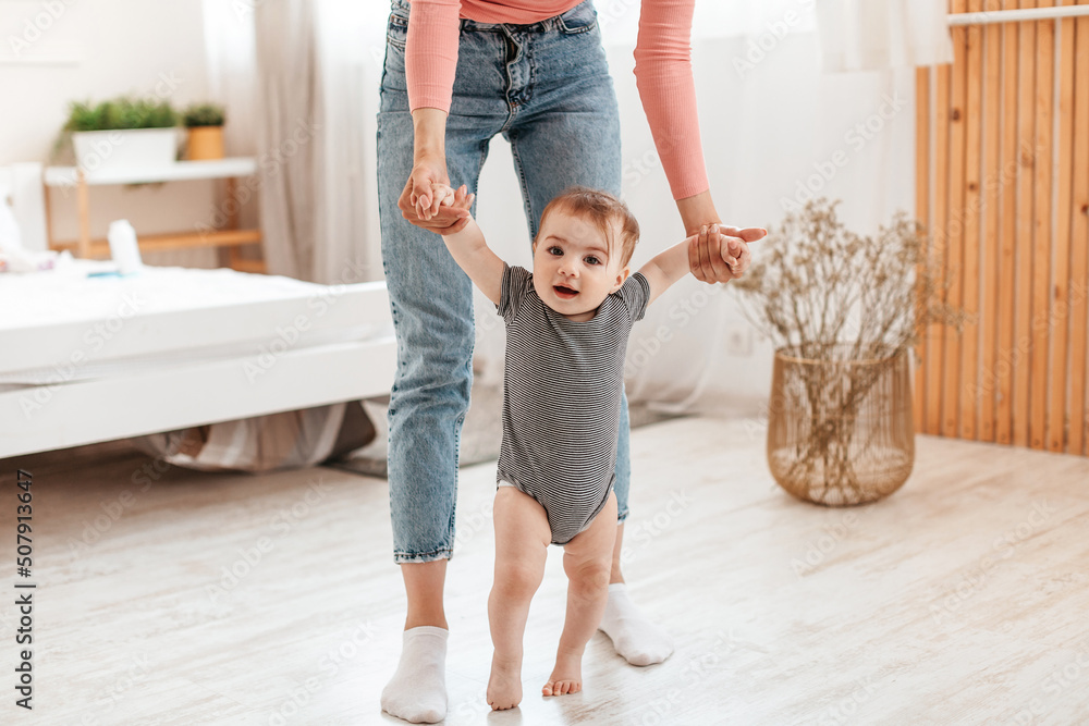 Adorable infant kid making first steps with mother's help, little baby holding mom's hands while walking in living room Adorable infant kid making first steps with mother's help, little baby holding mom's hands while walking in living room