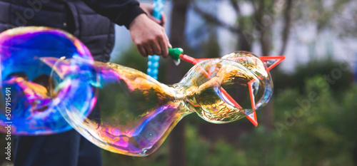 A man's hand makes big huge bright soap bubbles against the background of trees. Soap bubble show. Summer children's active recreation. Abstract blurred background.