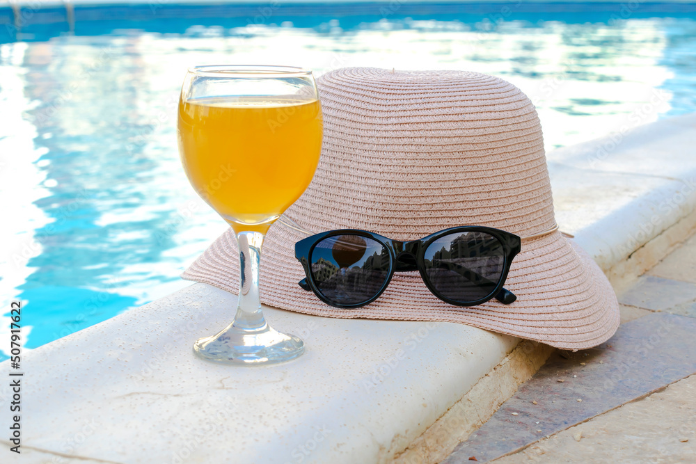 Closeup of yellow straw hat and black protective sunglasses, goggles, fresh orange juice smoothie drink cocktail on hotel swimming pool with blue water on warm sunny day. Summer vacation concept