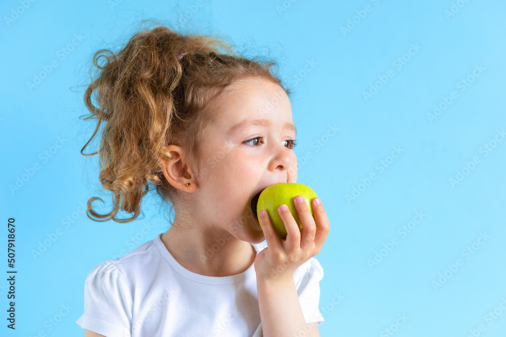 cute child smiling bites apple. nutrition for caries prevention. portrait of little girl with fruit on blue background. kid eating healthy food, snack. teeth, dental health, smile. children dentistry cute child smiling bites apple. nutrition for caries prevention. portrait of little girl with fruit on blue background. kid eating healthy food, snack. teeth, dental health, smile. children dentistry
