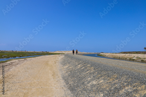 two women walking along a dirt footpath surrounded by vast blue ocean water and lush green grass with a gorgeous clear blue sky at Bolsa Chica Ecological Reserve in Huntington Beach California photo